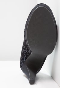 Chaussure à talon noir avec semelle texturée et dessus à motifs, photographiée par le dessous et en angle contre un mur et un sol blancs.