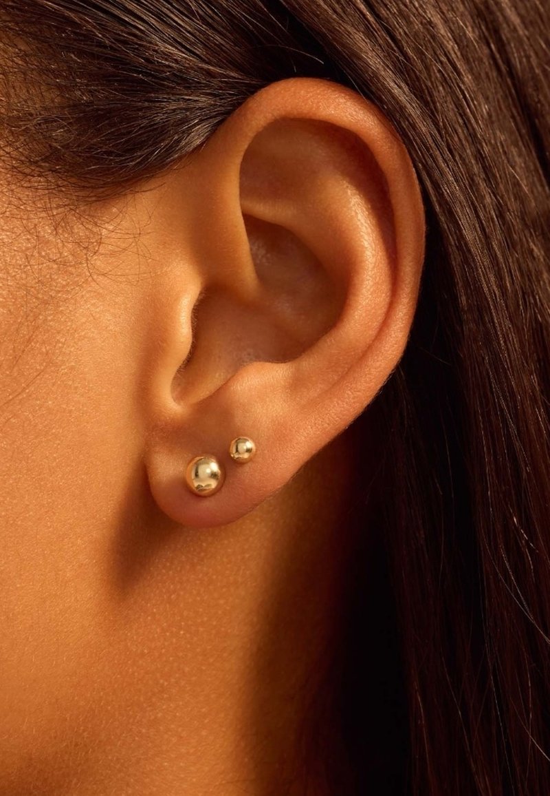 Boucles d'oreilles en or avec deux designs sphériques, un plus grand et un plus petit, fixées dans une oreille percée. Texture lisse avec des surfaces réfléchissantes.