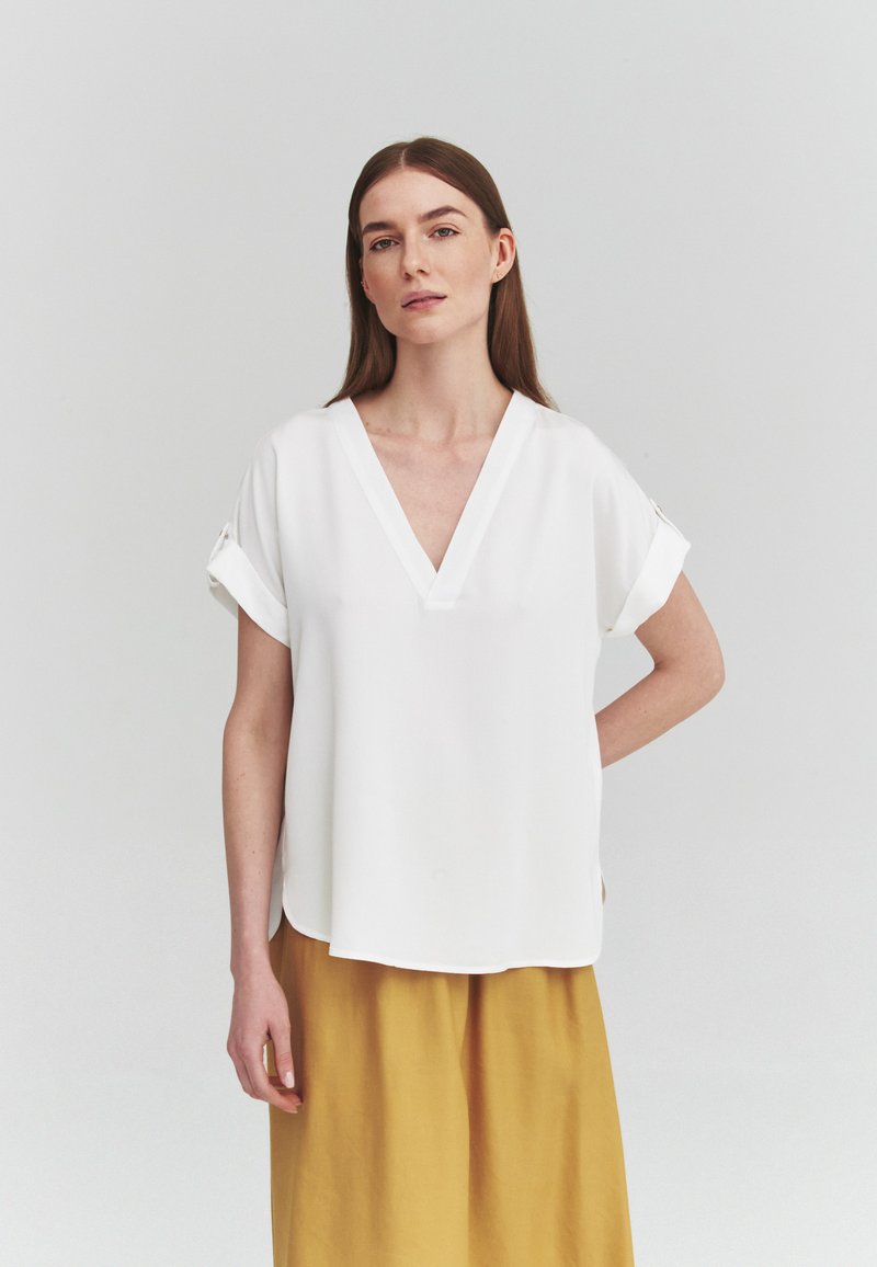 Femme aux longs cheveux bruns portant un chemisier blanc ample à col en V et une jupe jaune moutarde, debout devant un fond uni gris clair.