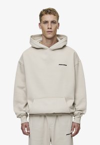 Beige hettegenser med kengurulomme, ribbede mansjetter og dristig svart "PEGADOR"-logo på brystet. Laget av børstet bomullsstoff.