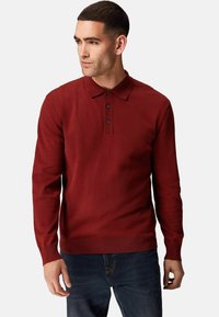 Rød langærmet polo sweater lavet af strikket stof. Har en knappeforespørgsel og ribbede manchetter. Kombineret med mørke denim jeans.