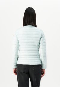 Vrouw met steil donker haar, gekleed in een lichtblauwe gewatteerde jas en zwarte jeans, gezien vanuit de achterkant tegen een effen witte achtergrond.