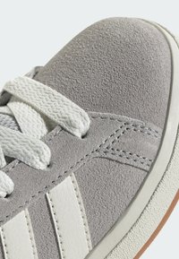 Šedé semišové tenisky s bílými koženými pruhy, texturovaným povrchem a bílými tkaničkami. Mají gumovou podešev a kontrastní švy.