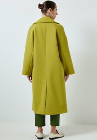 Long manteau vert surdimensionné avec un large col, des épaules tombantes et une fente à l'arrière, associé à un pantalon vert et des chaussures beige à enfiler.