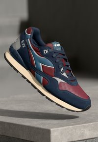 Navy and burgundy sneaker with a retro design, featuring prominent laces, mesh panels, and a contrasting sole, floating above a concrete surface.