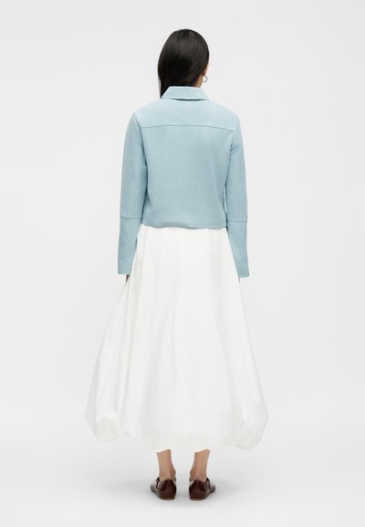 Moteris su ilgais tamsiais plaukais, dėvinti šviesiai mėlyną striukę, baltą ilgą sijoną ir rudus batus, stovinti nugara į žiūrovą, paprastoje fone.