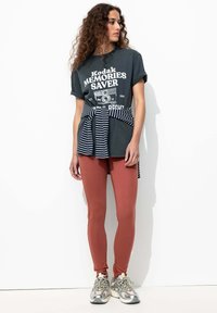 T-shirt gris foncé avec un graphique blanc "Kodak MEMORIES SAVER", tissu noué de rayures marines autour de la taille, associé à des leggings rouille et des baskets argentées.