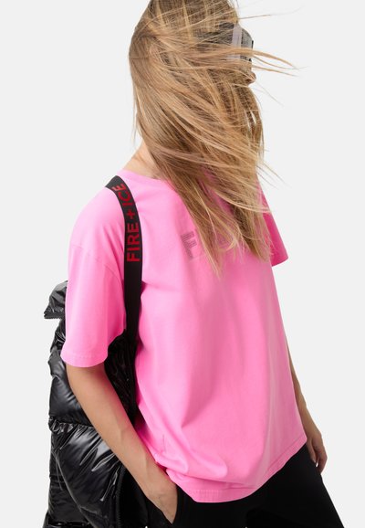 Person med langt hår, der blæser, iført en lys rosa t-shirt, sorte bukser og en sort jakke over den ene skulder med en "FIRE+ICE" rem.