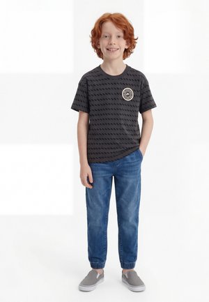 Uśmiechnięty rudowłosy chłopiec stoi z jedną ręką w kieszeni, ubrany w czarny T-shirt w wzory, niebieskie dżinsy i szare buty wsuwane na białym tle.
