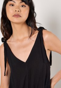 Black sleeveless dress with a deep V-neckline, featuring tied straps and a textured fabric. Fitted bodice leading to a relaxed skirt.