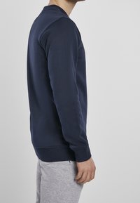 Felpa blu navy con scollo rotondo, maniche lunghe, texture morbida e polsini e orlo a coste. Design aderente, vista di profilo.