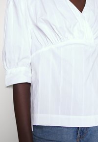 Blouse blanche à manches courtes en tissu texturé avec de fines rayures verticales, détail froncé à la taille et décolleté en V.