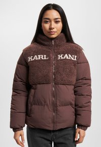 Brun pufferjakke med en fuzzy, struktureret overdel. Har en høj krave og broderet "KARL KANI" logo. Lynlåslukning og elastiske manchetter.