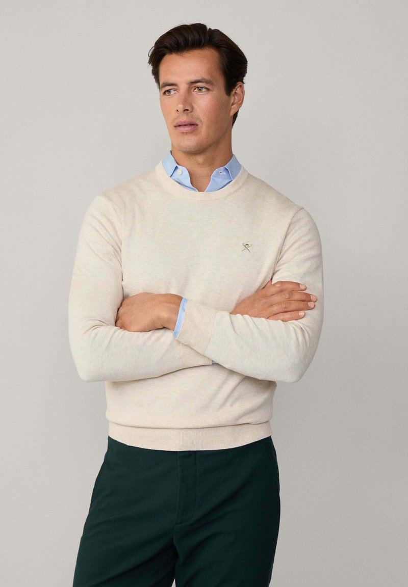 Beiger Pullover mit Rundhalsausschnitt, gerippten Bündchen und Saum. Mit kleinem besticktem Logo auf der Brust. Getragen über einem hellblauen Hemd.