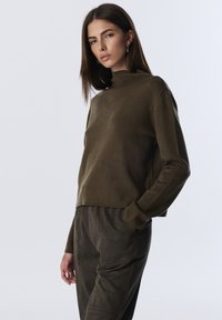 Młoda kobieta z długimi brązowymi włosami, ubrana w ciemnooliwkowy sweter z wysokim kołnierzem i dopasowane spodnie, pozująca z jedną ręką w kieszeni.