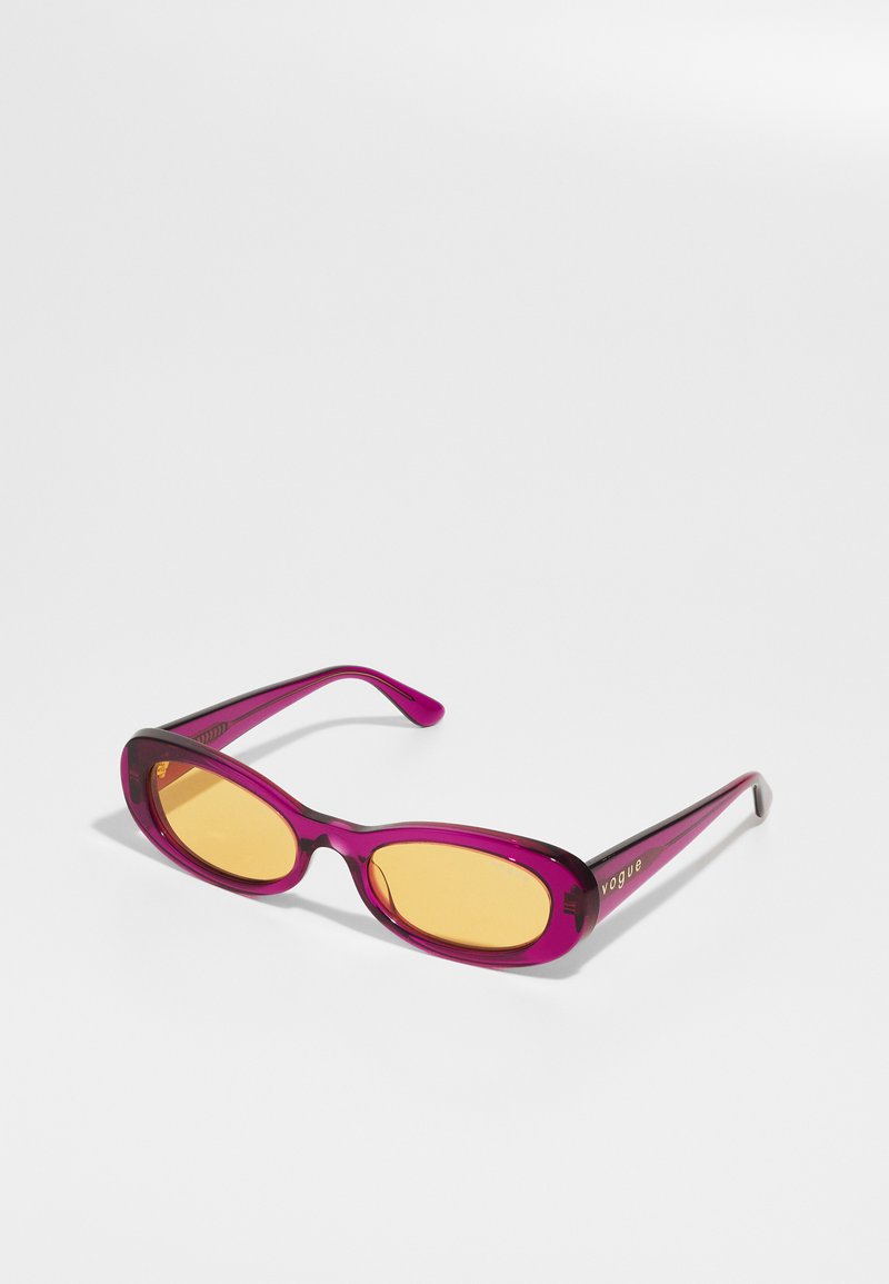 Pērļu caurspīdīgas ovālas saulesbrilles ar dzelteniem lēcām un "Vogue" uzrakstu zeltā uz kātu, novietotas uz baltas virsmas.