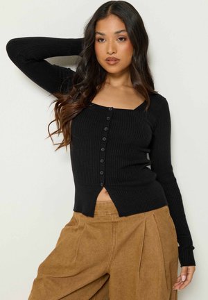 Femme aux longs cheveux foncés portant un pull côtelé noir boutonné et un pantalon large marron taille haute, posant avec un bras derrière la tête.