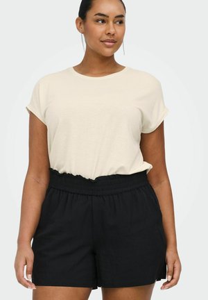 Kvinde med mørkt hår i en knold iført en beige kortærmet top, der er stoppet ned i sorte højtaljede shorts med en flæsekant ved taljen.