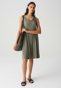 Olive green sleeveless dress with a V-neck, flowing silhouette, and soft fabric. Black woven bag and flat black sandals are also shown.