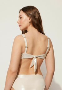 Světle béžový bikiny top s saténovou texturou, nastavitelnými ramínky a zavazováním na zadní straně. Jednoduchý design s minimálním množstvím kovových akcentů.