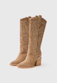 Bottes de cowboy en daim tan avec des clous en argent, des motifs brodés, des pointes effilées, des languettes de tirage et des talons blocs empilés.