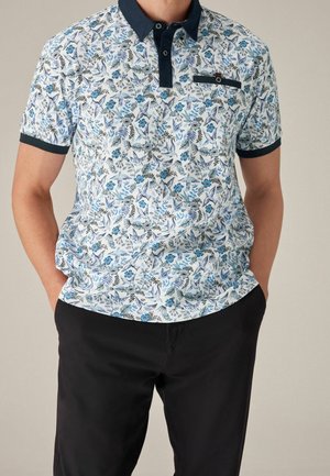 Homme portant un polo blanc à manches courtes avec des motifs floraux et d'oiseaux bleus, col noir, et pantalon noir, mains dans les poches.