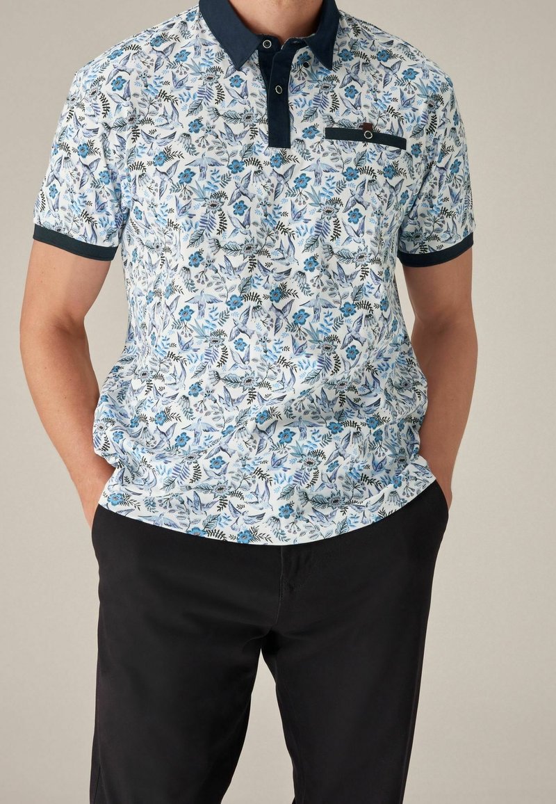 Homme portant un polo blanc à manches courtes avec des motifs floraux et d'oiseaux bleus, col noir, et pantalon noir, mains dans les poches.