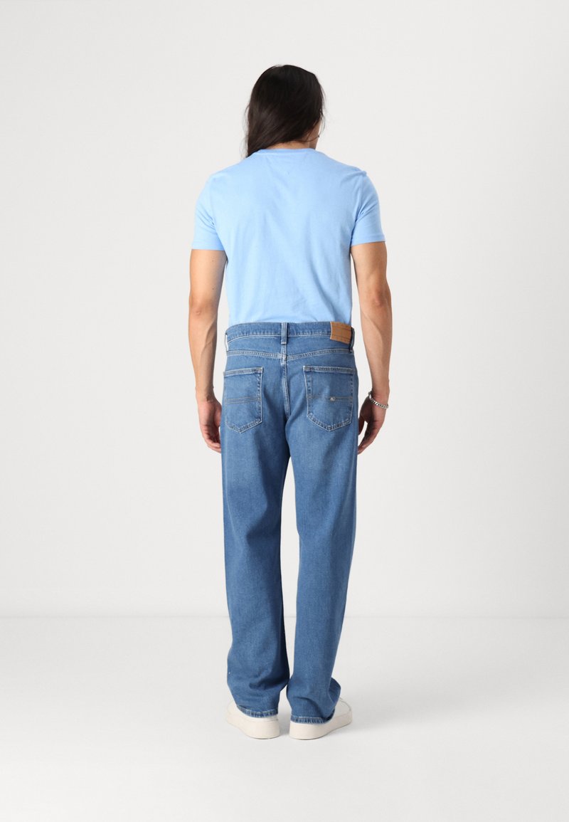 Helles blaues T-Shirt und lockere blaue Denim-Jeans mit einem Lederpatch am hinteren Bund und weißen Sneakern. Schlichte Gestaltung.