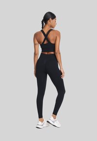 Zwarte sportbeha met kruislings straps, gecombineerd met hoge zwarte leggings. Beide gemaakt van een gladde, rekbare stof. Witte sneakers maken de look compleet.