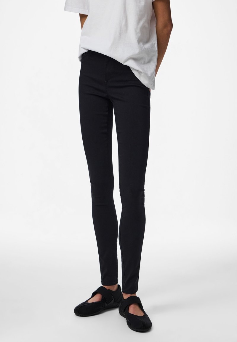Personne portant un pantalon noir moulant, un t-shirt blanc ample et des chaussures noires sans lacets avec des bandes Velcro, debout sur un fond blanc.