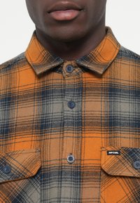 Camisa de franela a cuadros en naranja, gris y azul marino. Presenta cierres de botones y dos bolsillos frontales con etiqueta de marca. Textura suave.