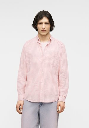 Personne aux cheveux châtains mi-longs portant une chemise à carreaux rose clair à boutons et un jean bleu clair, debout devant un fond uni.