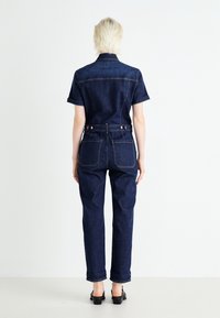 Combinaison en denim foncé à manches courtes, coupe ajustée, coutures apparentes et deux poches arrière. Les détails comprennent des passants de ceinture et des boutons en métal.