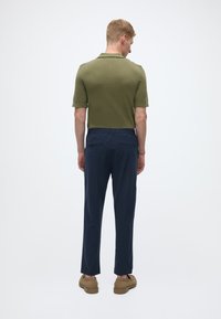 Polo vert olive à manches courtes associé à un pantalon bleu marine. Chaussures marron. Vue de dos mettant en valeur un design simple et des couleurs unies.