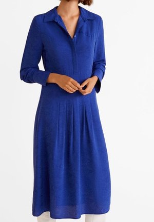 Vrouw die een midi-jurk met lange mouwen, kraag en plooien vanaf de taille omlaag draagt, handen losjes vooraan samengevouwen.