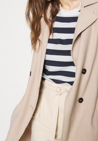 Béžový kabát s vystřiženým límcem a tmavými knoflíky, přes námořnickou a bílou pruhovanou košili, zkombinován se světle béžovými kalhotami s páskem zavázaným v pase.