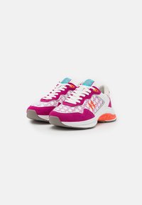 Valkoiset ja magentanväriset sneakersit, joissa oranssit tehosteet, siksak-kuvio, valkoiset nauhat ja turkoosit tunnuslaput valkoista taustaa vasten.