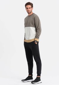 Herreoutfittet inkluderer en tofarvet sweatshirt i oliven og creme, med beige detaljer, parret med sorte tapered joggers og sorte løbesko.