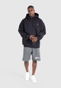 Felpa nera in cotone con tasca a marsupio frontale, indossata con pantaloni sportivi grigi con logo, abbinati a sneakers bianche.