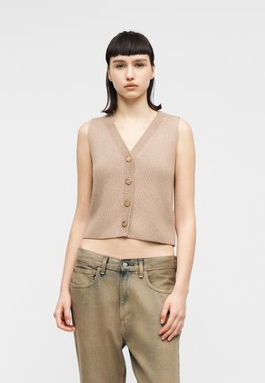 Rövid barna hajú nő, aki bézs színű, ujjatlan, gombos kötött mellényt és laza, kifakult barna farmert visel, egyszínű háttér előtt áll.