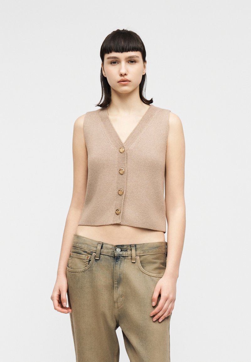 Γυναίκα με κοντά σκούρα μαλλιά φορώντας γιλέκο από πλεκτό ύφασμα χωρίς μανίκια σε μπεζ χρώμα με κουμπιά και χαλαρά ξεθωριασμένα καφέ τζιν, στέκεται μπροστά σε ένα απλό φόντο.