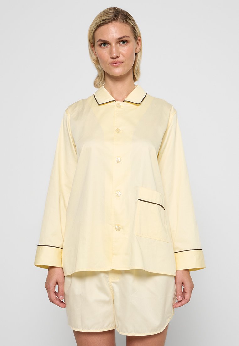 Conjunto de pijama de algodón amarillo con mangas cortas, camisa abotonada que presenta un bolsillo y ribetes en negro, junto con pantalones cortos a juego.