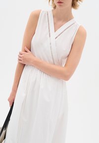 Abito bianco senza maniche con design a portafoglio, caratterizzato da un bordo testurizzato vicino al collo e una vita arricciata. La modella tiene una borsa nera.