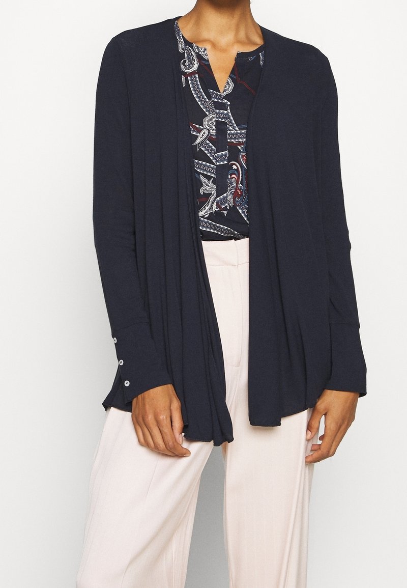 Granatowy kardigan z otwartym przodem, długimi rękawami i guzikami na mankietach, noszony na wzorzystej bluzce i jasnych spodniach.