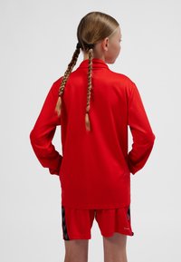 Maglia atletica rossa a maniche lunghe, abbinata a pantaloncini coordinati con accenti a chevron neri. Tessuto liscio ed elastico.