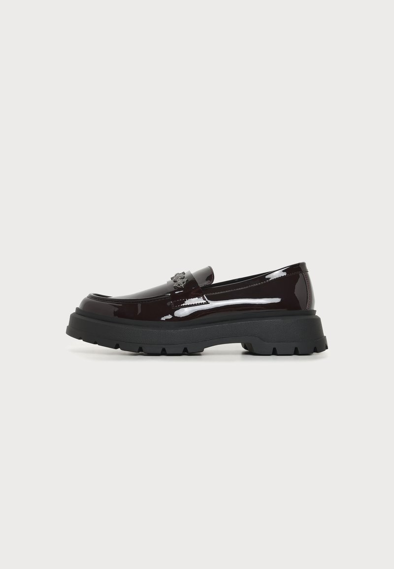 Mocassin marron foncé brillant avec une semelle noire épaisse et un petit emblème décoratif sur le dessus, présenté en profil latéral sur un fond uni.