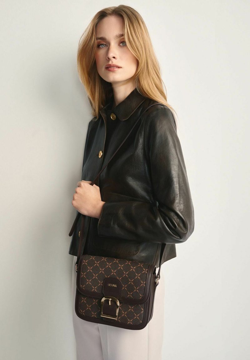 Młoda kobieta z blond włosami nosi czarną skórzaną kurtkę i beżowe spodnie, trzymając ciemnobrązową torebkę na ramię w wzór z złotą klamrą.