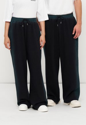 Zwei Personen tragen schwarze Hosen mit weitem Bein und Kordelzug in der Taille sowie weiße Sneaker und stehen nebeneinander vor einem weißen Hintergrund.