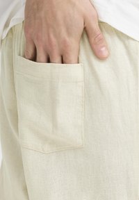 Lyse beige linnedbukser med et enkelt design, der har en enkelt påsyet lomme på højre side. Glat tekstur og afslappet pasform.