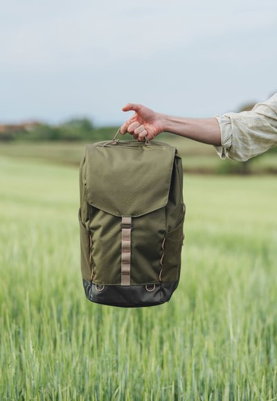 Olivgrön ryggsäck med en vikbar lucka, brunt accentband, sidofickor och en svart bottenpanel. Tillverkad av slitstarkt tyg, utrustad med ett handtag.